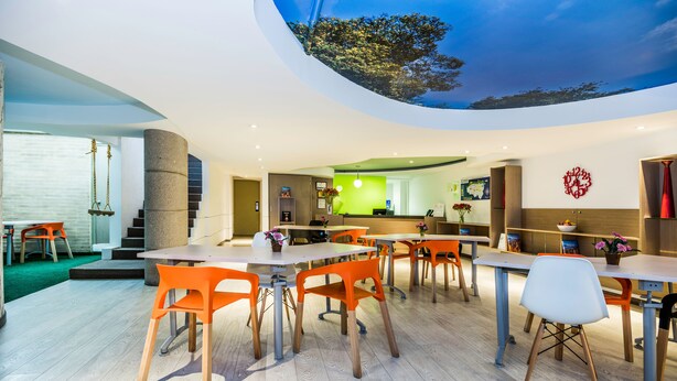 A modern interior with a dining area featuring wooden tables and orange chairs, a reception desk, and a large mural on the ceiling. - Viaggio Virrey (Bogotá)