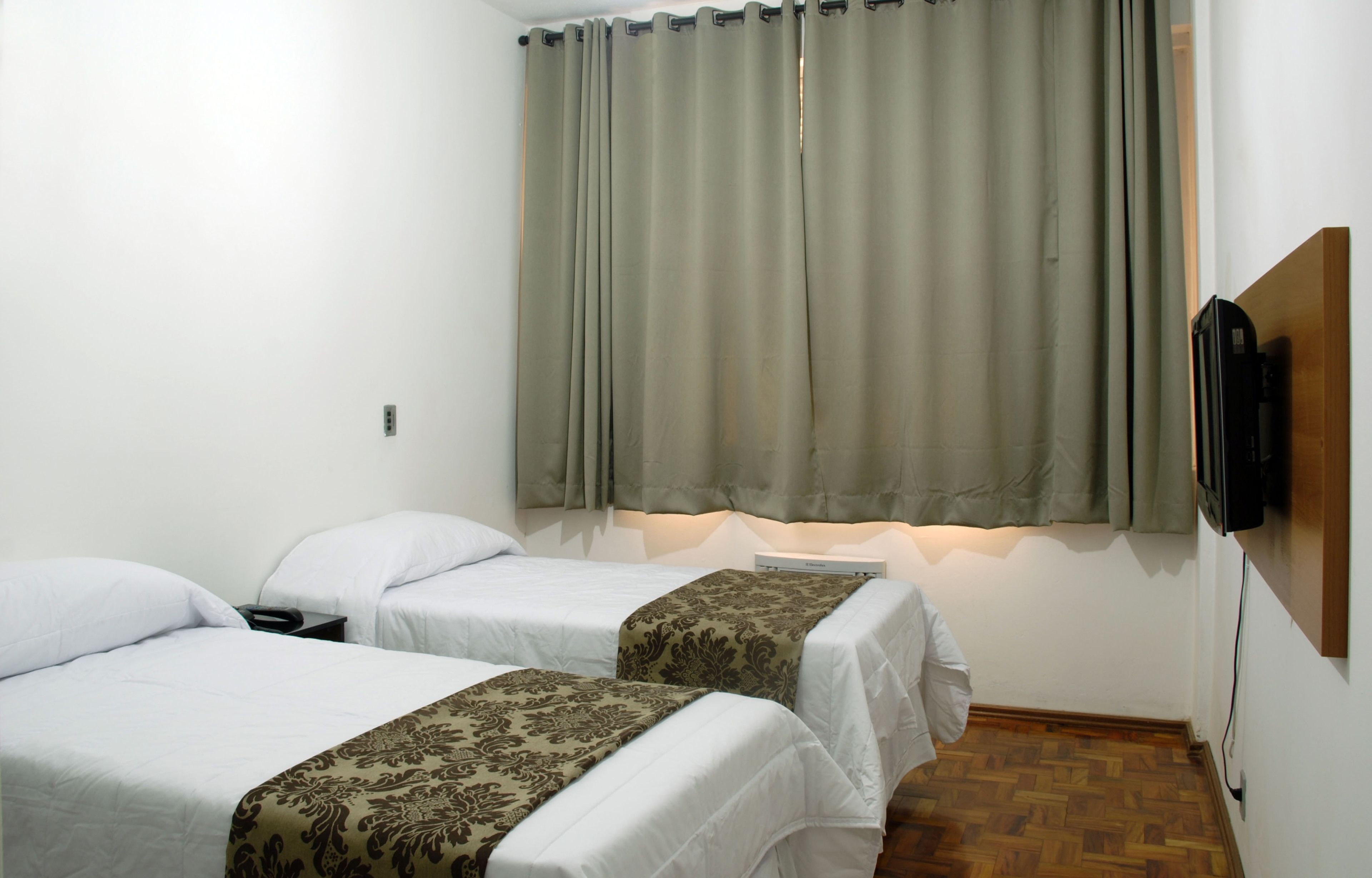 A room with two single beds, a flat-screen TV mounted on the wall, and a window with curtains.