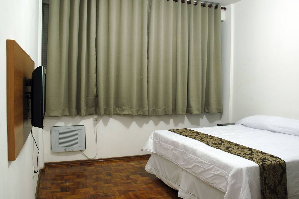 Ett hotellrum med en säng, en tv som är monterad på väggen, en värmare och ett fönster med gardiner.