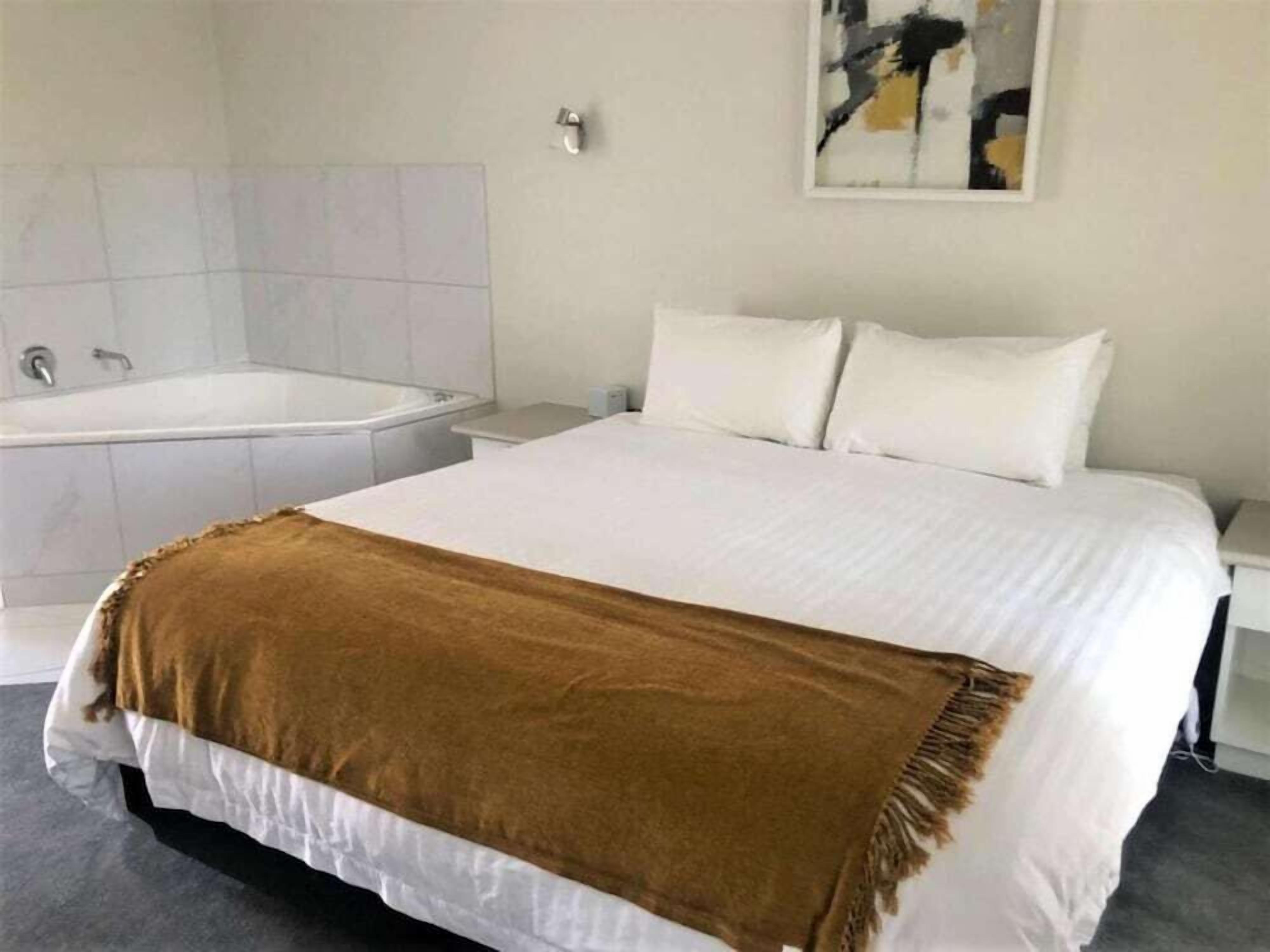 A bedroom with a large bed, a white headboard, a brown throw blanket, a white nightstand, and a white wall with a painting.
