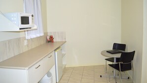 Uma pequena cozinha compacta com micro-ondas, pia e bancada. Há uma mesa pequena com duas cadeiras e uma janela com cortinas.