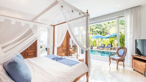 A bedroom with a four-poster bed, a television, and a view of a pool and outdoor seating. - AQ-VA Hotel & Villas Seminyak (Legian)