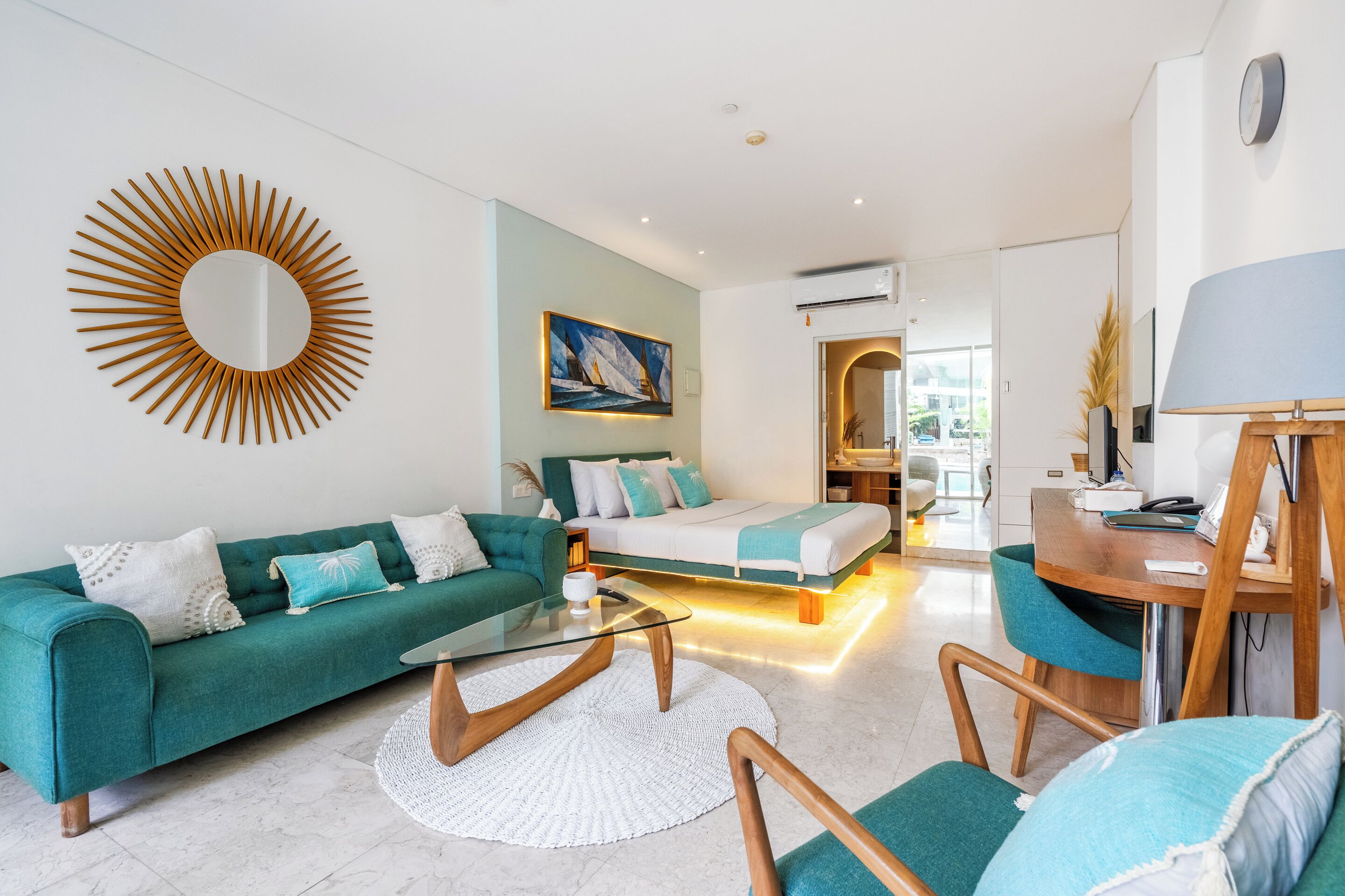 A modern living room with a teal sofa, a glass coffee table, and a wooden accent chair. A bed with a white and teal duvet is visible in the background.