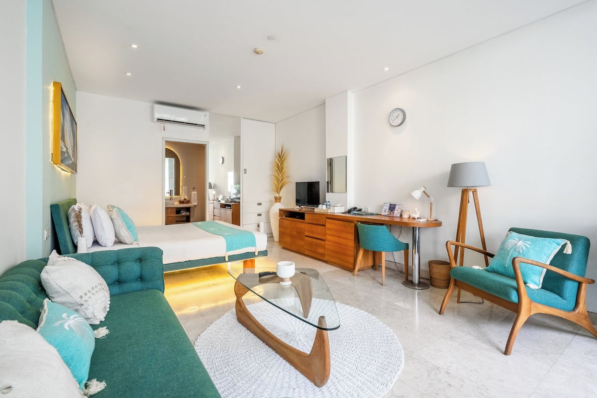 Ein modernes Wohnzimmer mit einer Couch, einem Couchtisch, einem Schreibtisch und einem Sessel. Im Hintergrund ist ein Bett zu sehen, sowie ein Esstisch mit Stühlen.