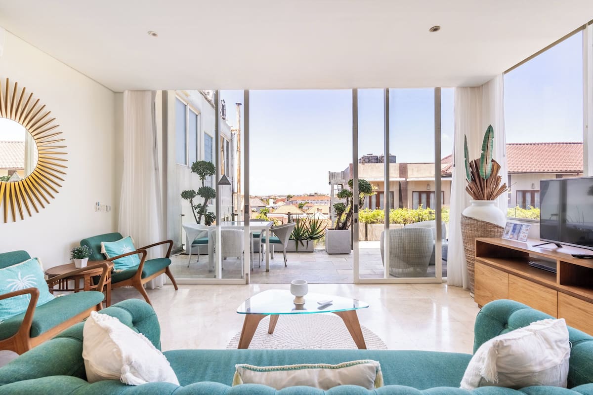 Ein geräumiges Wohnzimmer mit einem großen Fernseher, grünen Sofas, einem Glastisch und Blick auf die Stadt durch große Fenster.