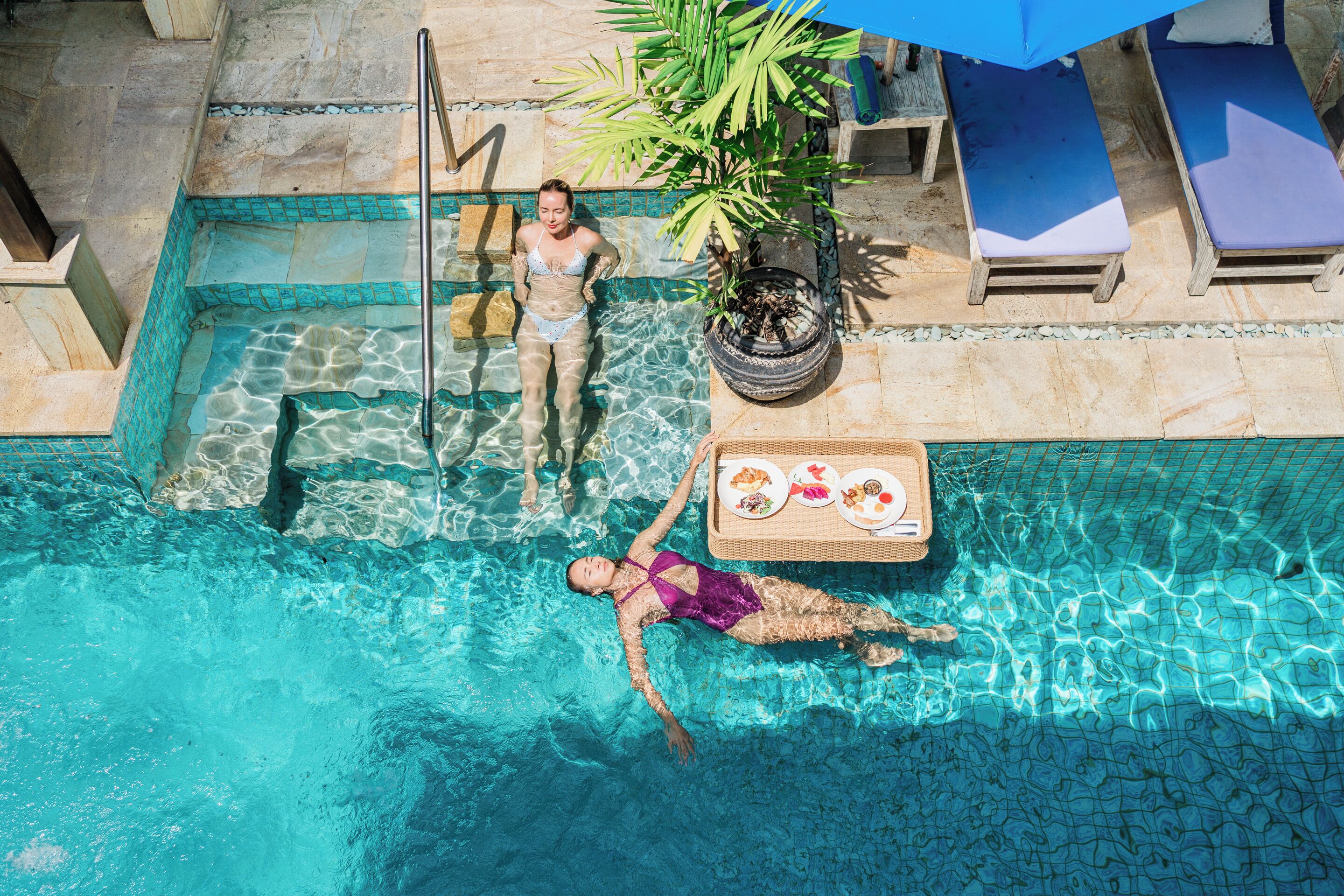 A swimming pool with clear water, lounge chairs, and a dining table with food.