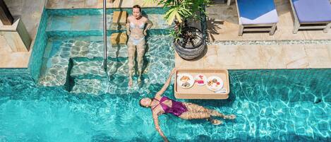 A swimming pool with clear water, lounge chairs, and a dining table with food.