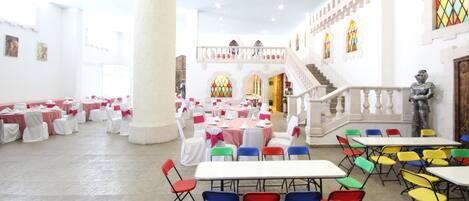 A banquet hall with tables set for events, a central column, and a staircase leading to an upper level.