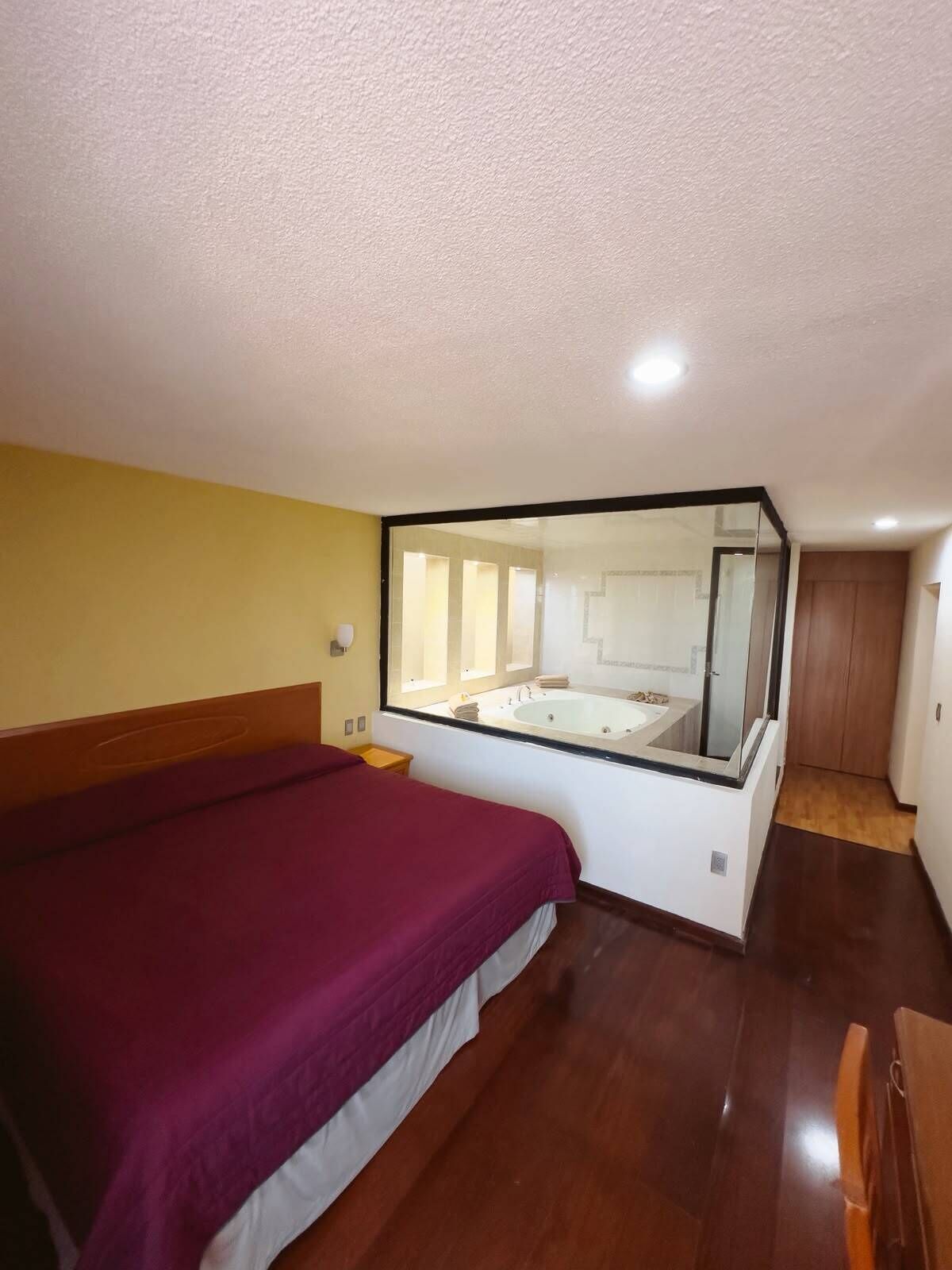 Ein Schlafzimmer mit einem großen Bett, einem durch eine Glaswand abgetrennten Badezimmer mit einer Badewanne und einem Holzboden.