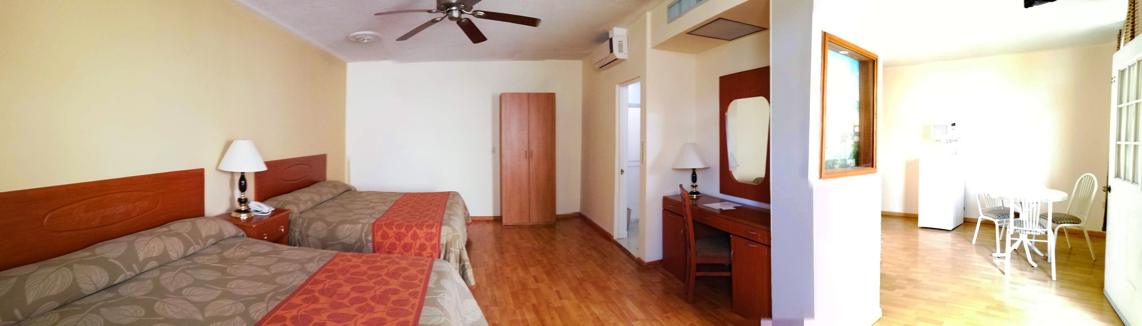 A hotel room with two beds, a desk, a chair, a mirror, and a ceiling fan.