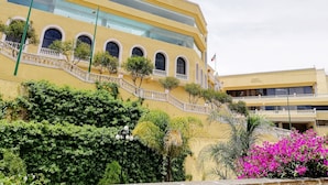 Un edificio amarillo con múltiples ventanas y balcones, rodeado de vegetación y una escalera.