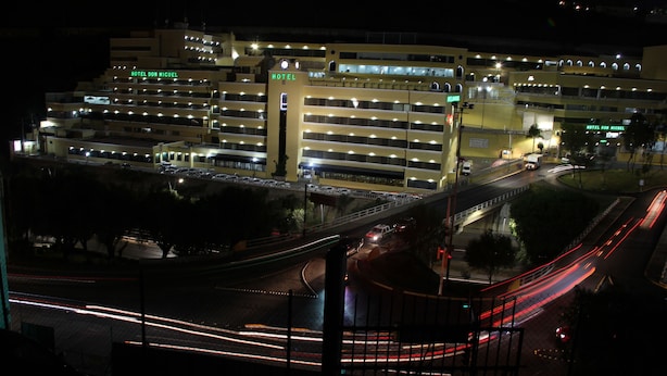 Un hotel de varios pisos con señalización iluminada, un estacionamiento y una calle concurrida con estelas de luz.