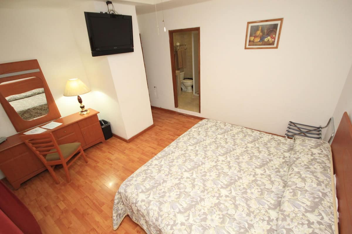 Quarto com cama, escrivaninha e cadeira, televisão e porta para o banheiro.