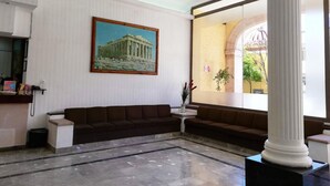 A lobby with a large painting of a classical building, a brown sofa, a small table with a vase, and a large column. - Hotel María Benita (Zacatecas)