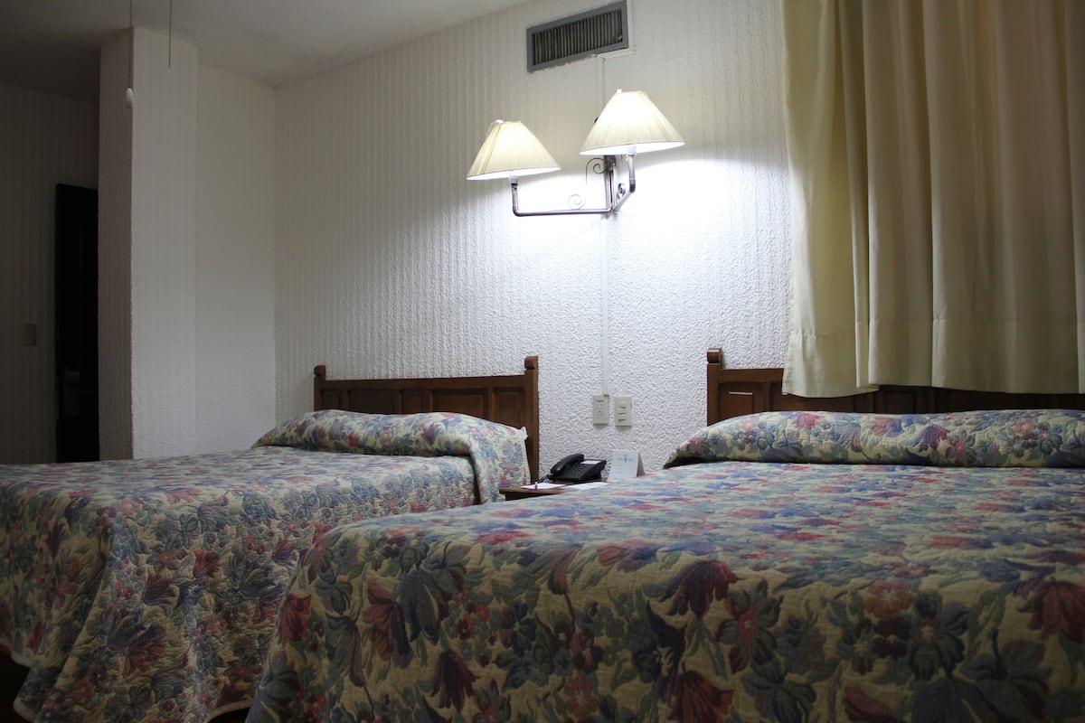 Quarto de hotel com duas camas, colchas estampadas com flores, um telefone e duas luminárias fixadas na parede.