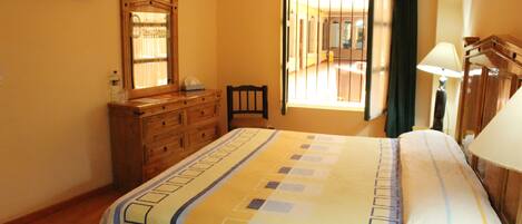 Quarto de hotel com cama, cómoda de madeira, espelho, janela com cortinas e televisão montada na parede.