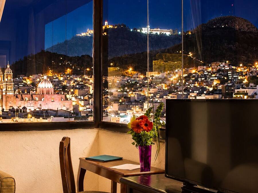 Una stanza con un'ampia finestra vista sulla città di notte, una televisione, una scrivania con una sedia e un vaso con fiori.