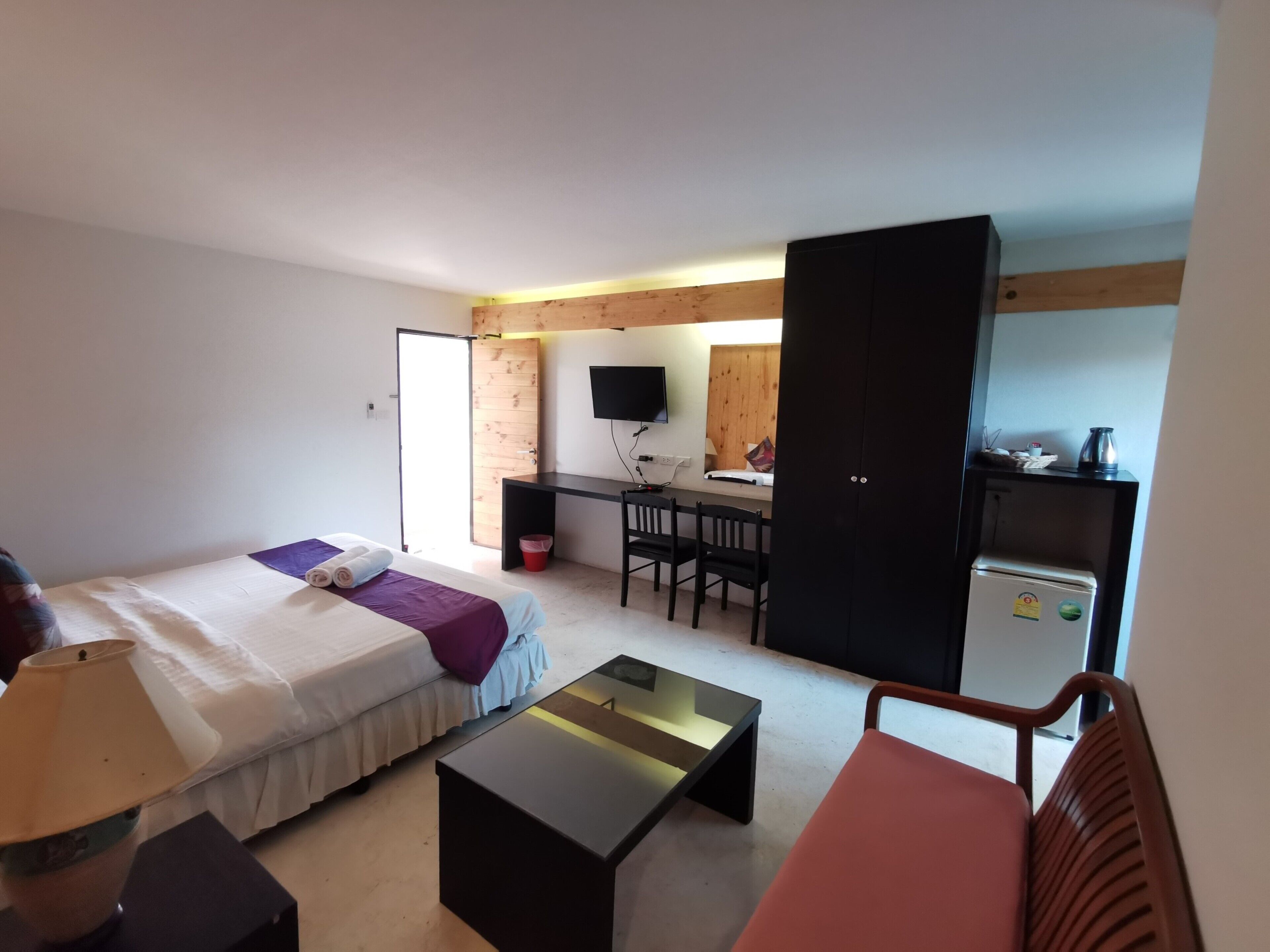 A compact hotel room with a bed, a desk, a chair, a small table, a TV, and a kitchenette.