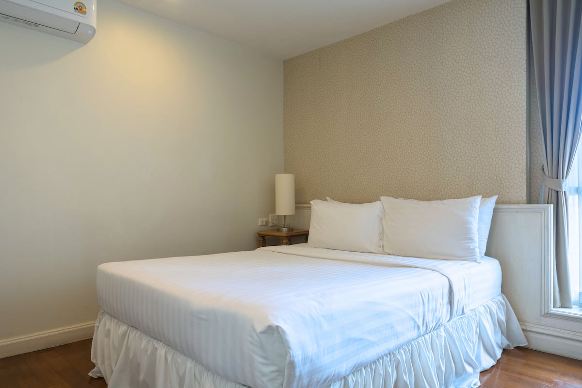 A neatly made bed in a hotel room with a plain wall, a nightstand with a lamp, and a window with curtains.