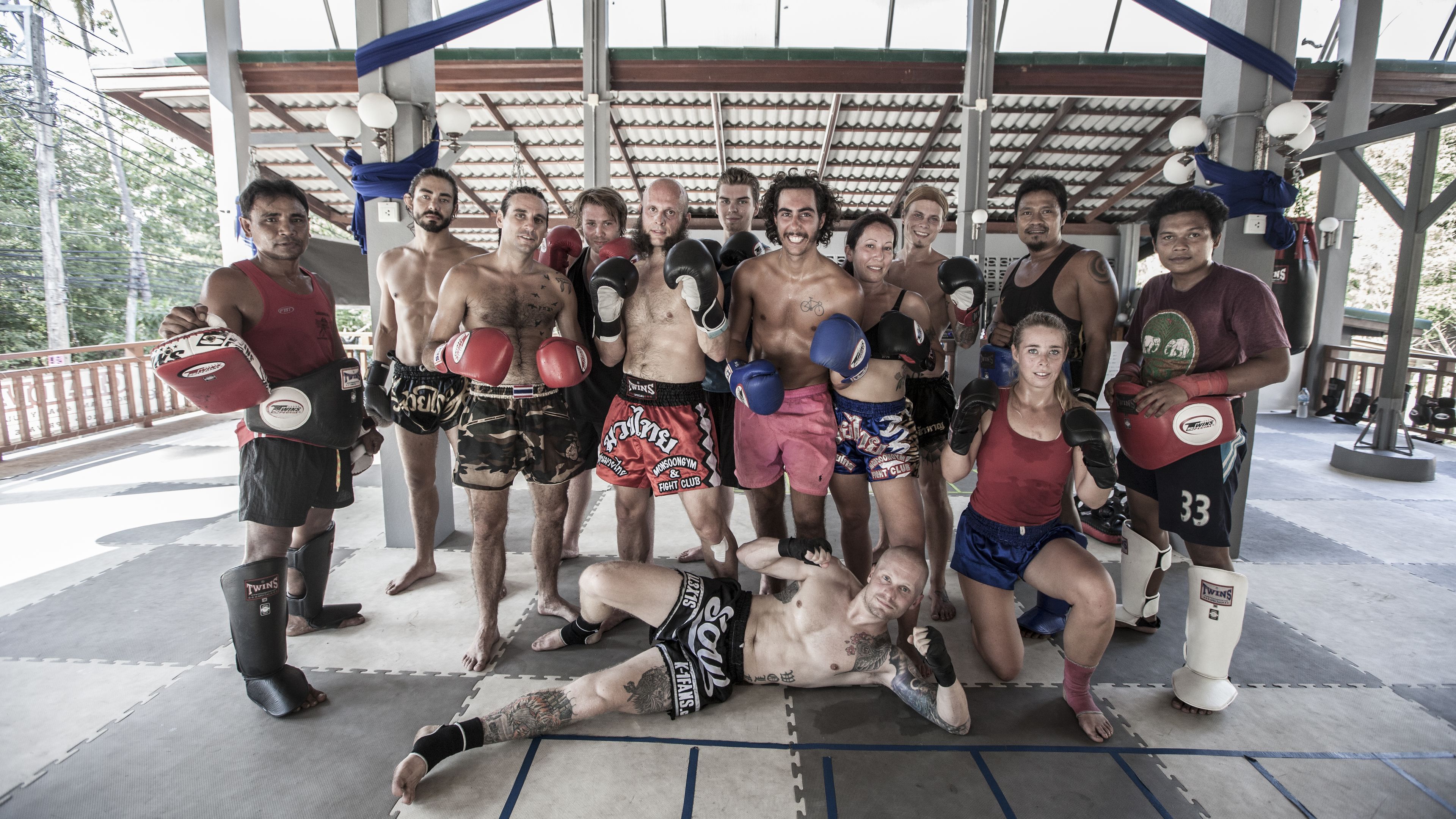 A group of people in a covered outdoor area with a roof, mats on the floor, and boxing gloves.