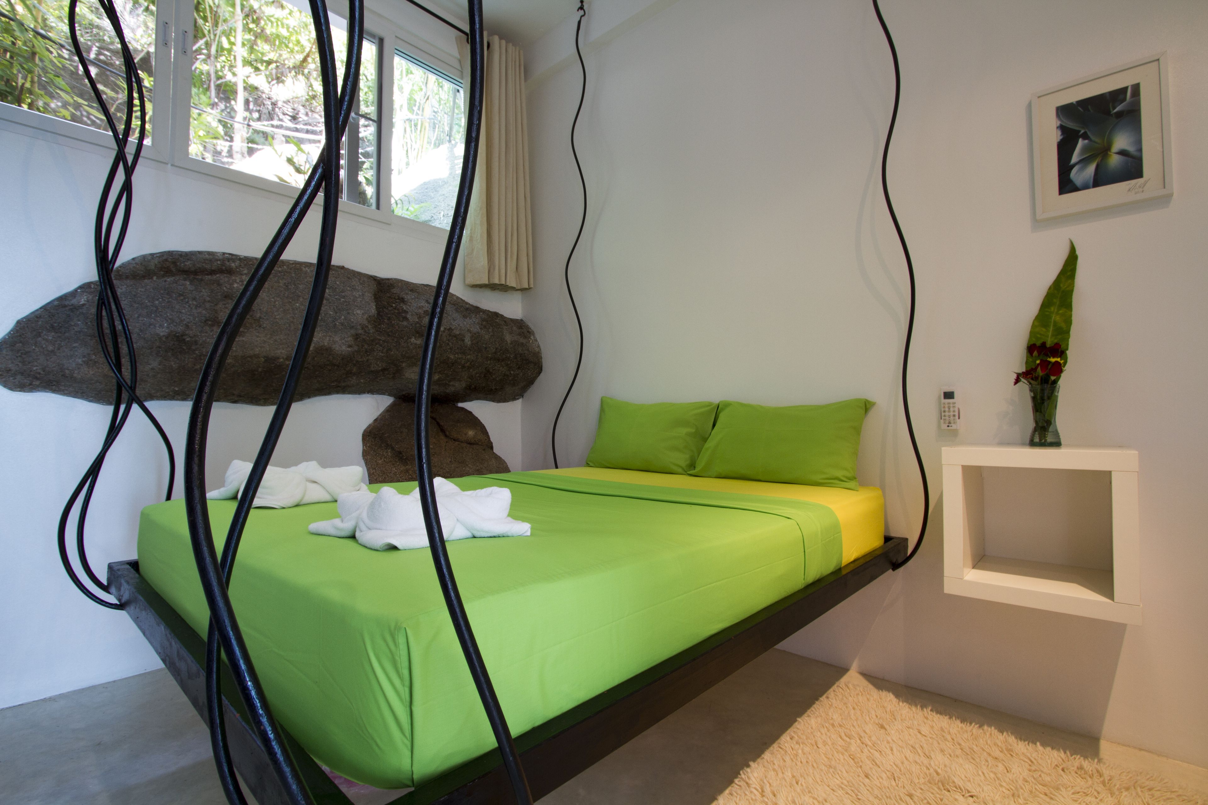 A modern bedroom with a hanging bed, a green mattress, a white pillow, and a small bedside table with a vase of flowers.