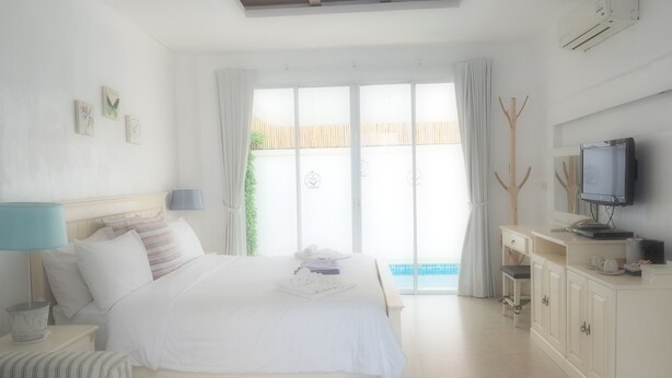 A bedroom with a bed, bedside table, lamp, TV, and a view of a pool. - Costa Village Pool Villa (Pattaya)
