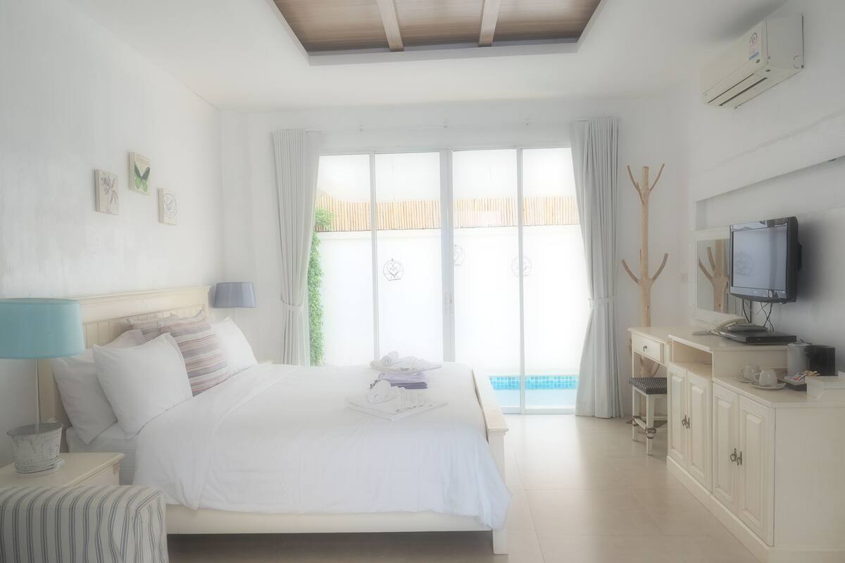 A bedroom with a bed, bedside table, lamp, TV, and a view of a pool.