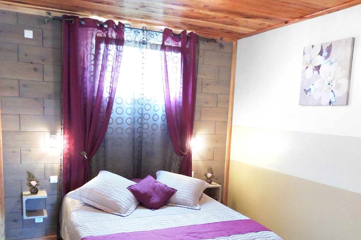 Un dormitorio con techo de madera, cama con ropa de cama blanca y detalles morados, una mesita de noche con un jarrón y una pared con un cuadro floral.