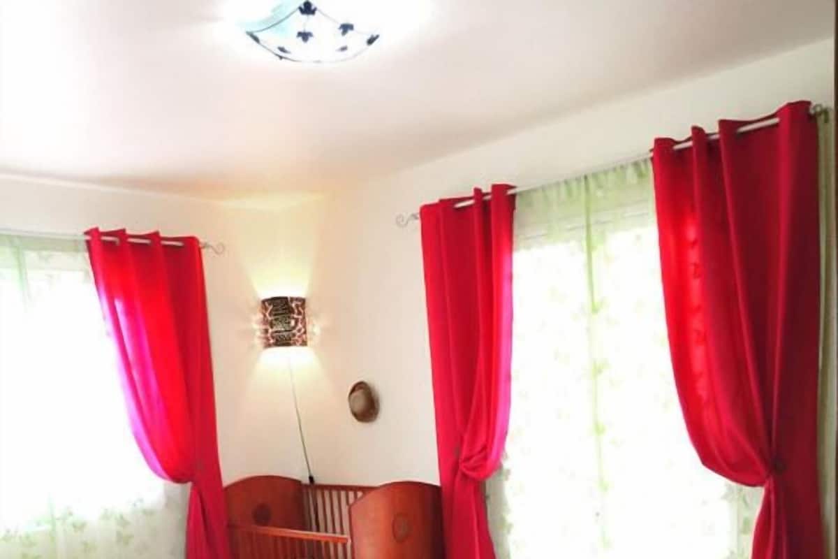 Un dormitorio con cama de madera, cortinas rojas, alfombra verde y una lámpara fijada a la pared.