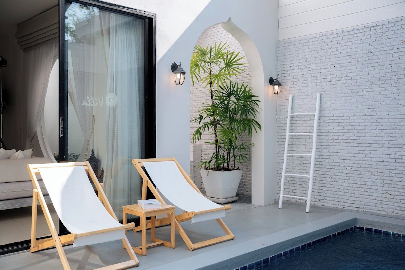A poolside area with two lounge chairs, a small table, a potted plant, and a white ladder.
