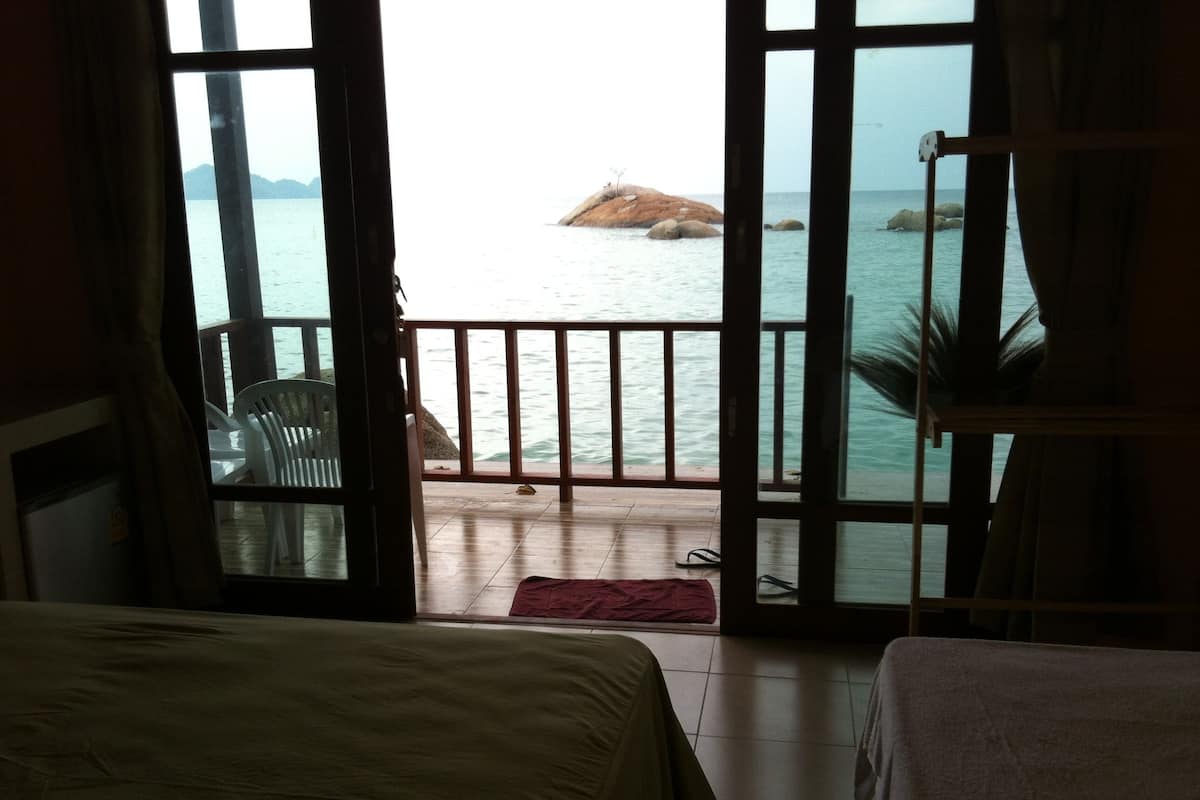 Una habitación con una cama, una mesita y una silla. Se ve el mar a través de una ventana con barandilla de madera.