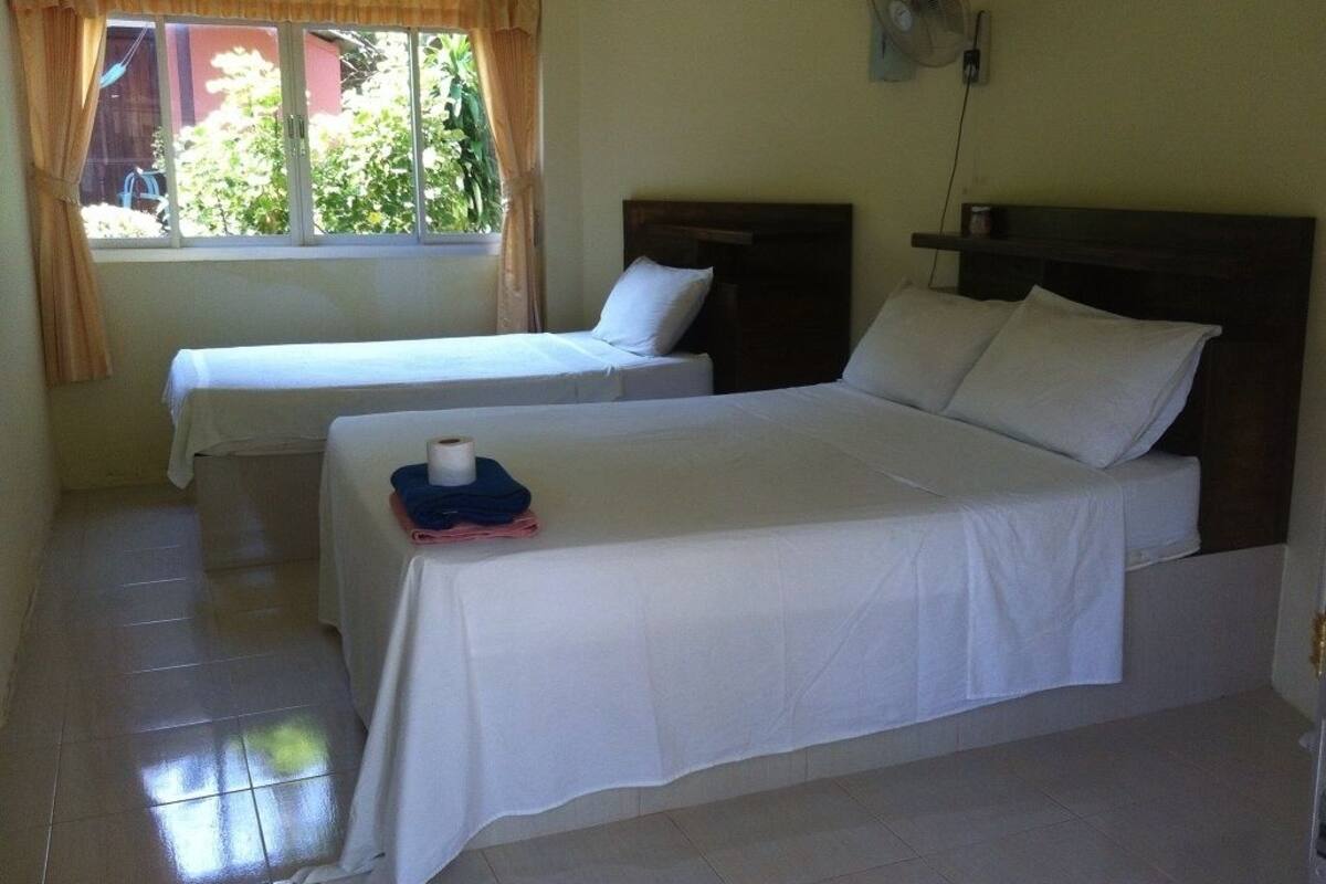 Una habitación con dos camas, una ventana con cortinas, un ventilador en la pared y una mesa con una toalla y un objeto azul.