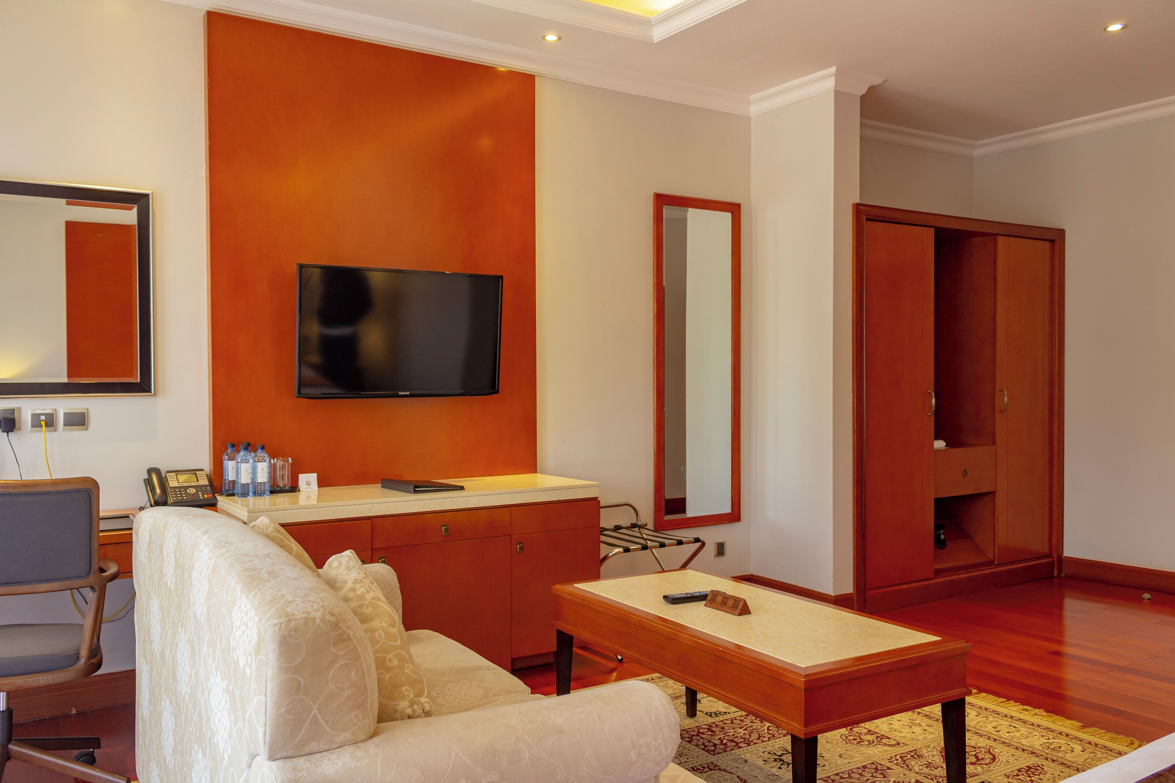 Une chambre d’hôtel équipée d’un téléviseur à écran plat fixé au mur, d’une coiffeuse en bois avec un miroir, d’un canapé, d’une petite table et d’une armoire.