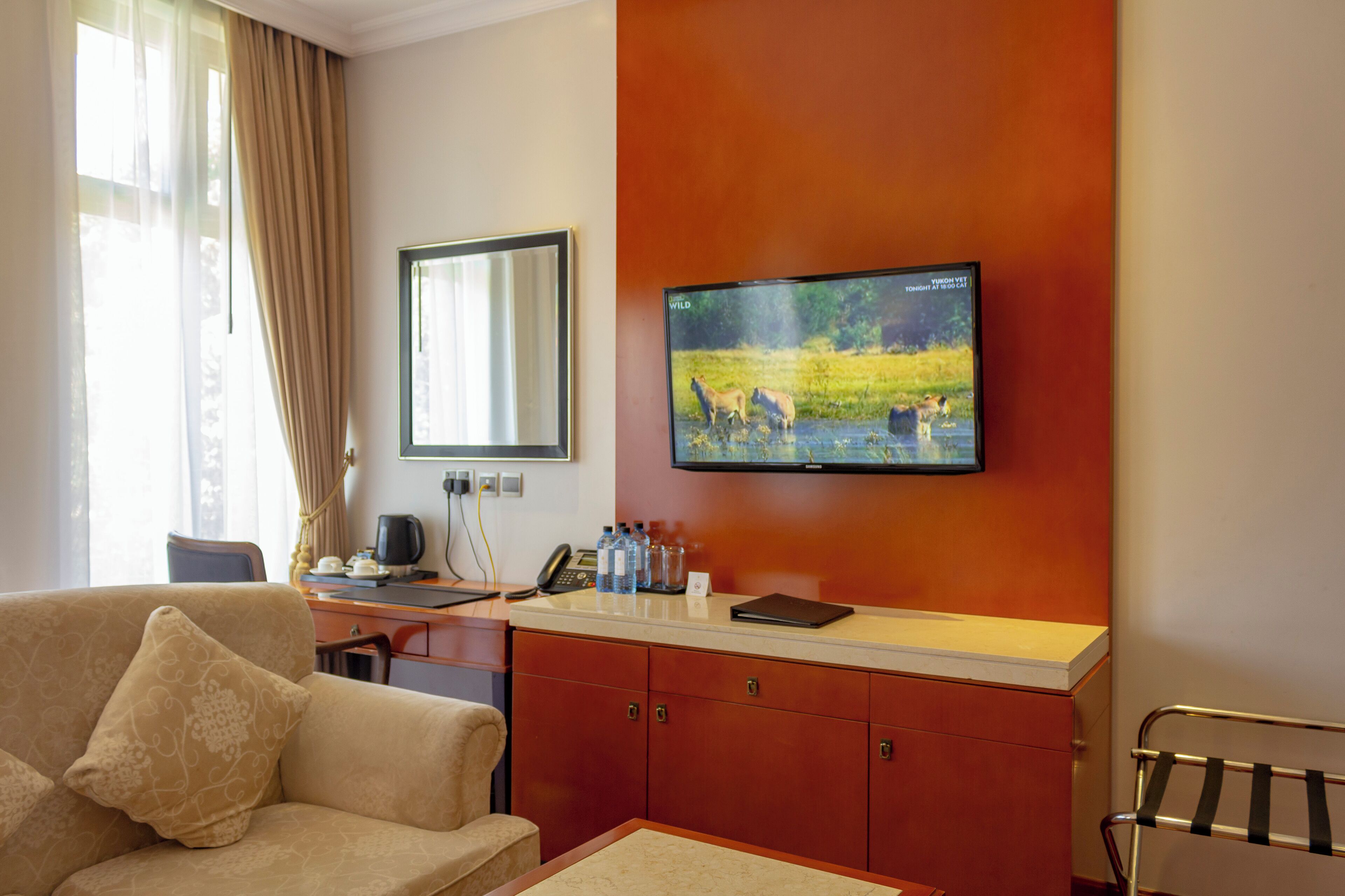 A hotel room with a sofa, a desk with a computer and telephone, a television mounted on the wall, and a window with curtains.