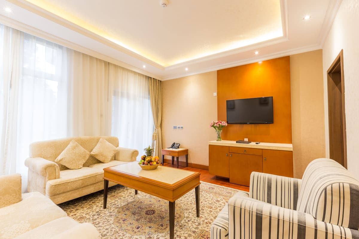 A well-lit living room with a sofa, armchair, and a coffee table. A flat-screen TV is mounted on the wall, and a small side table with a vase of flowers is visible.