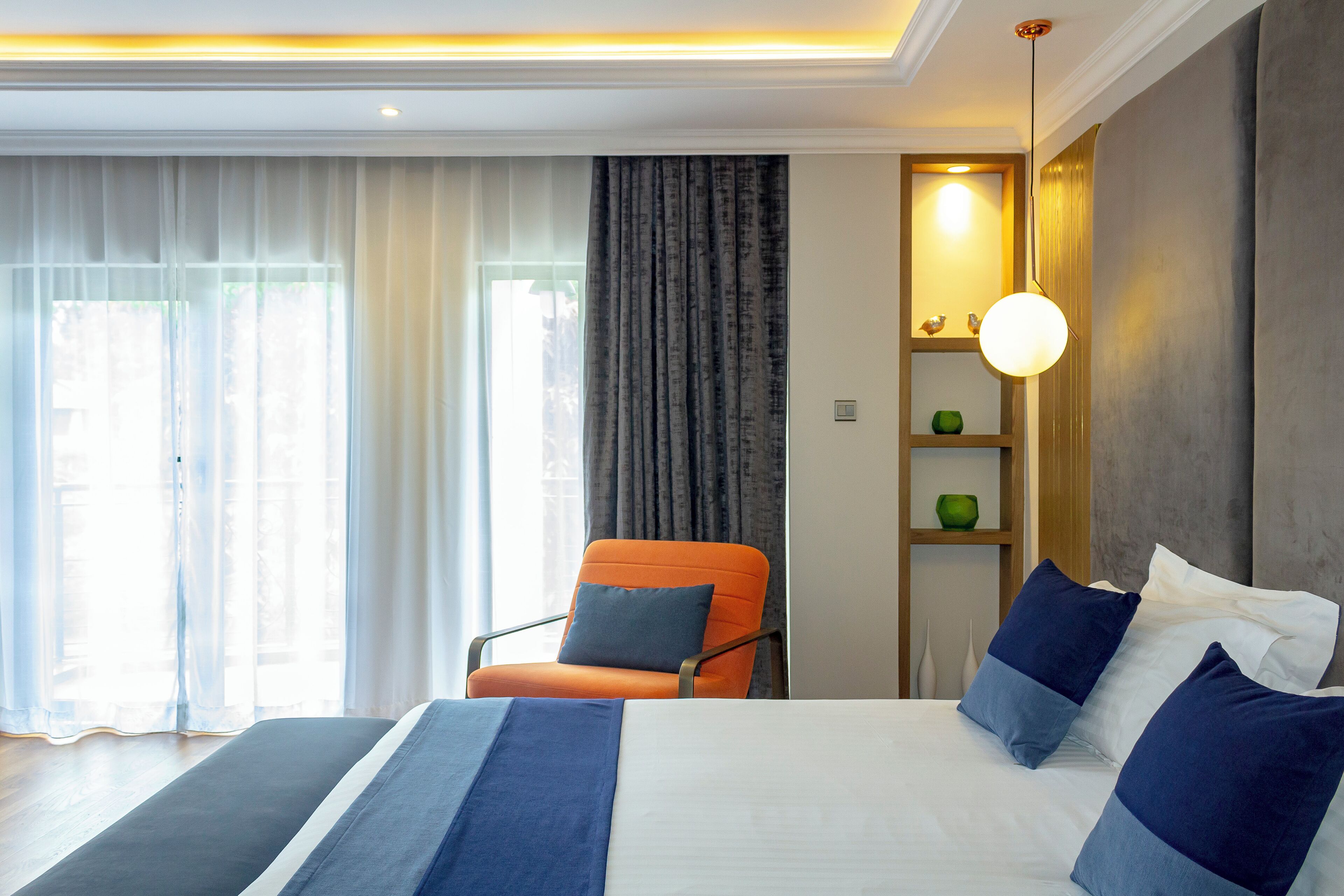 A modern hotel room with a large bed, an orange chair, a bedside table with a lamp, and a view of the outdoors through sheer curtains.