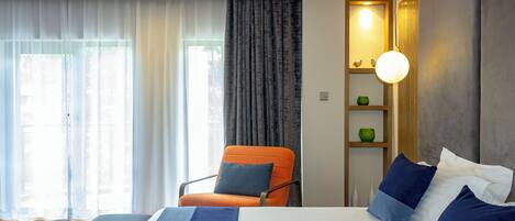 A modern hotel room with a large bed, an orange chair, a bedside table with a lamp, and a view of the outdoors through sheer curtains.