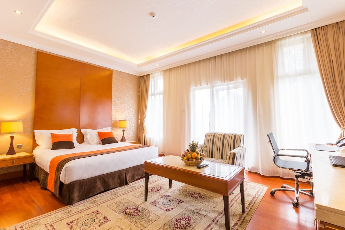 A hotel room with a large bed, a desk with a chair, a small table with a fruit bowl, and a window with curtains.