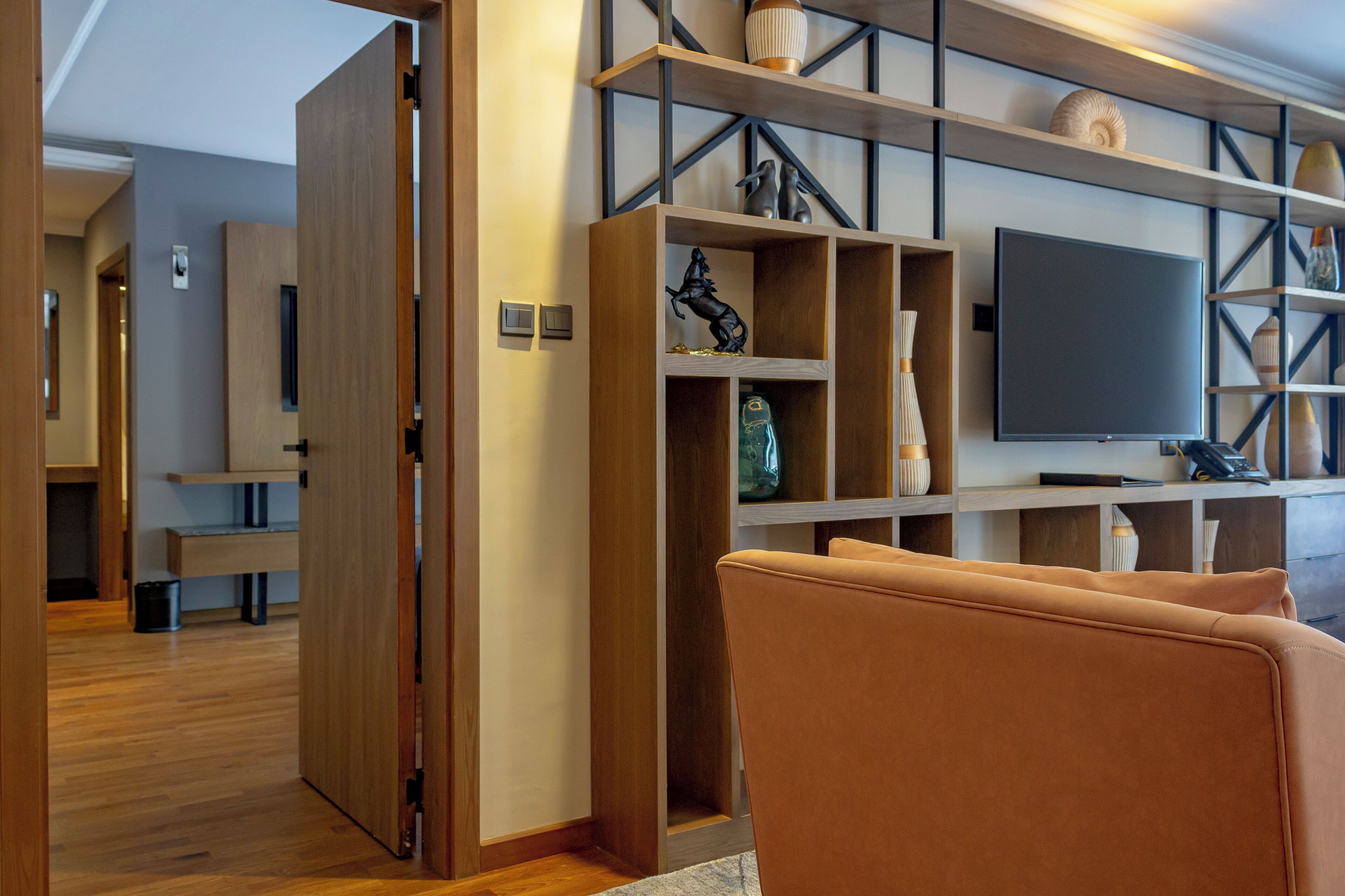 A modern living room with a wooden floor, a sofa, a TV mounted on a wall with shelves above, and a door leading to another room.