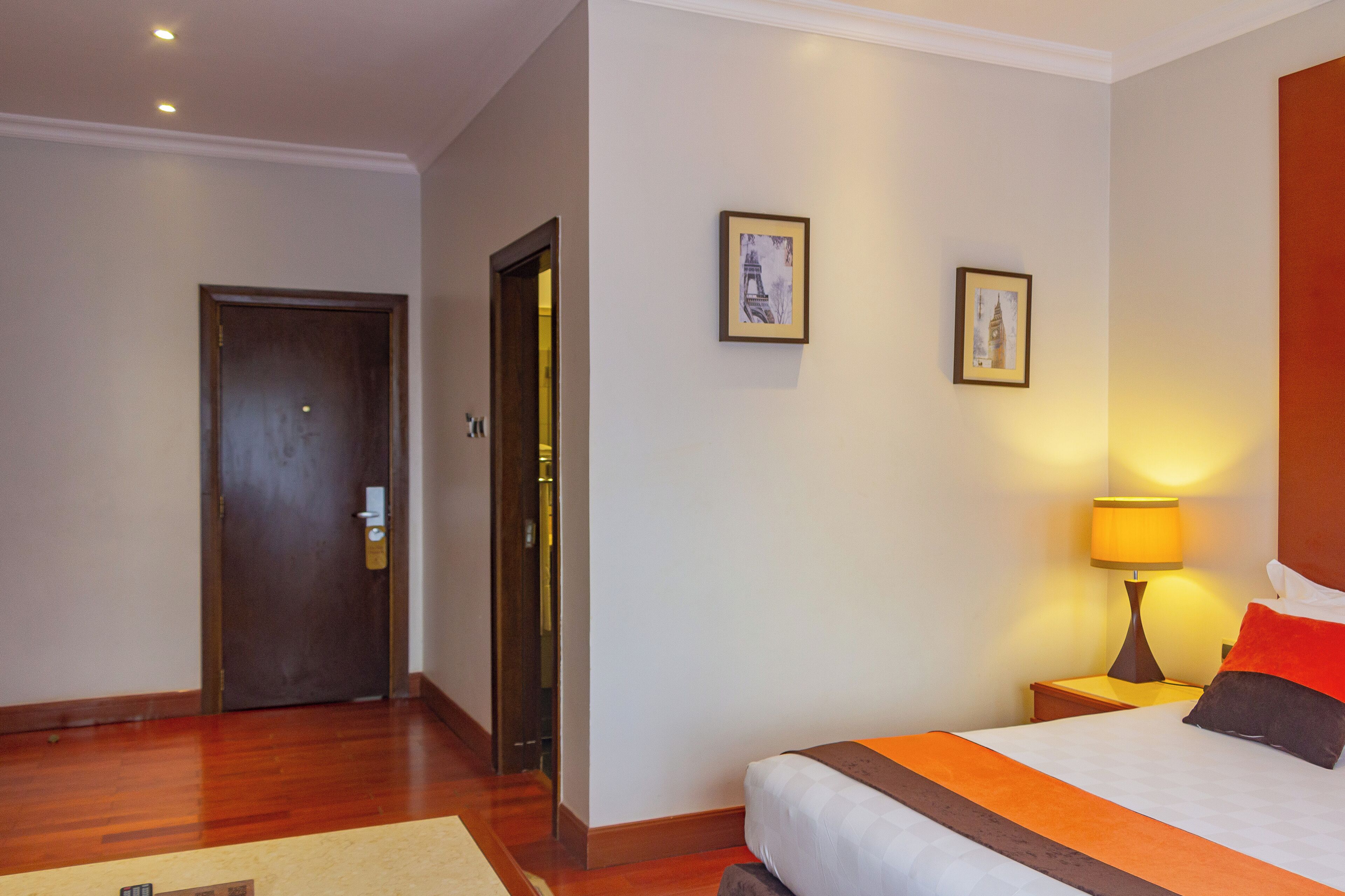 A hotel room with a bed, a nightstand with a lamp, a door, and two framed pictures on the wall.