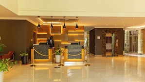 A hotel reception area with a modern design, featuring a reception desk, a waiting area with chairs, and a glass elevator. - Alba Hotel Meru (Meru)