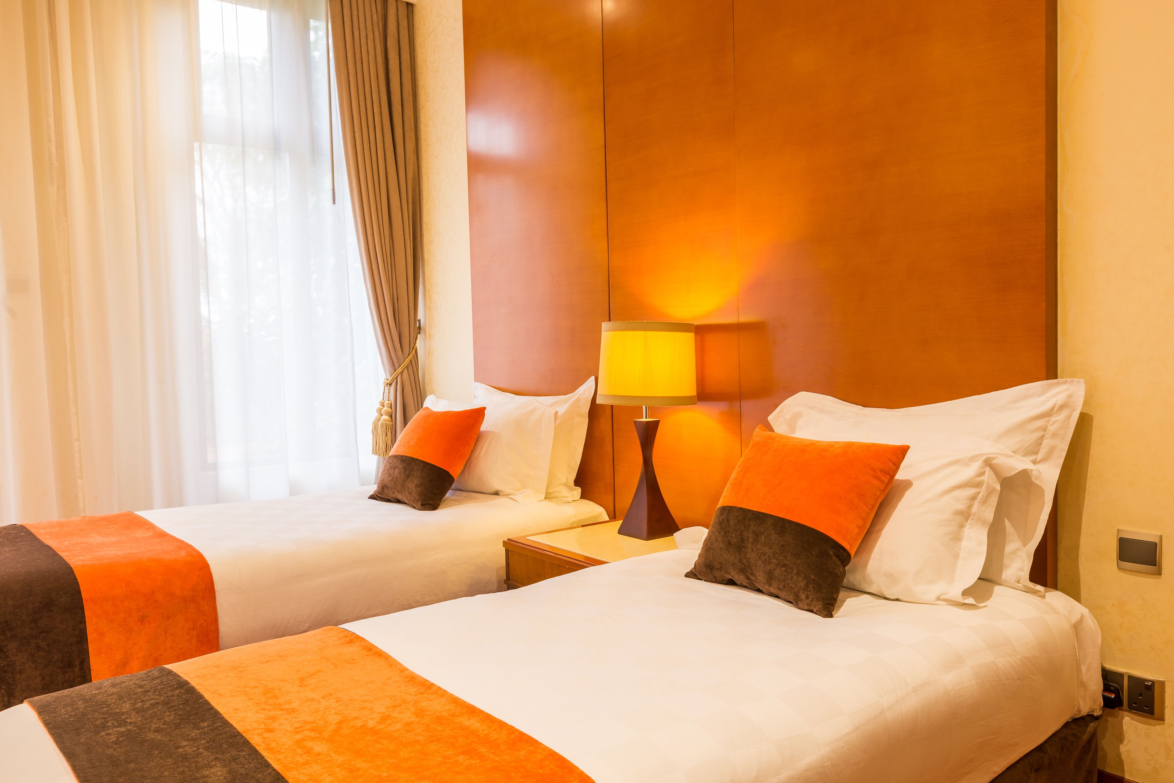 Een hotelkamer met twee bedden, elk met wit en oranje beddengoed, een nachtkastje met een lamp en een houten hoofdbord.