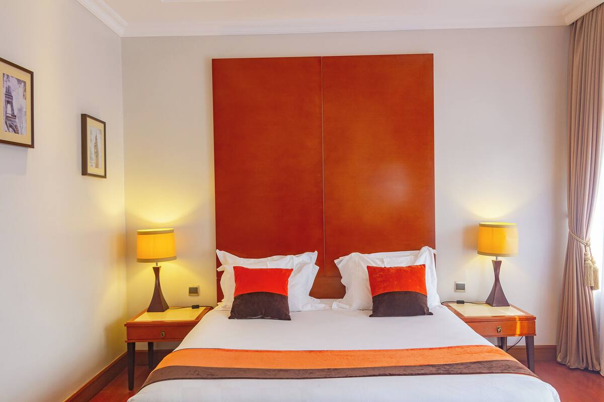 A hotel room with a large bed, two bedside tables with lamps, a wooden headboard, and a window with curtains.