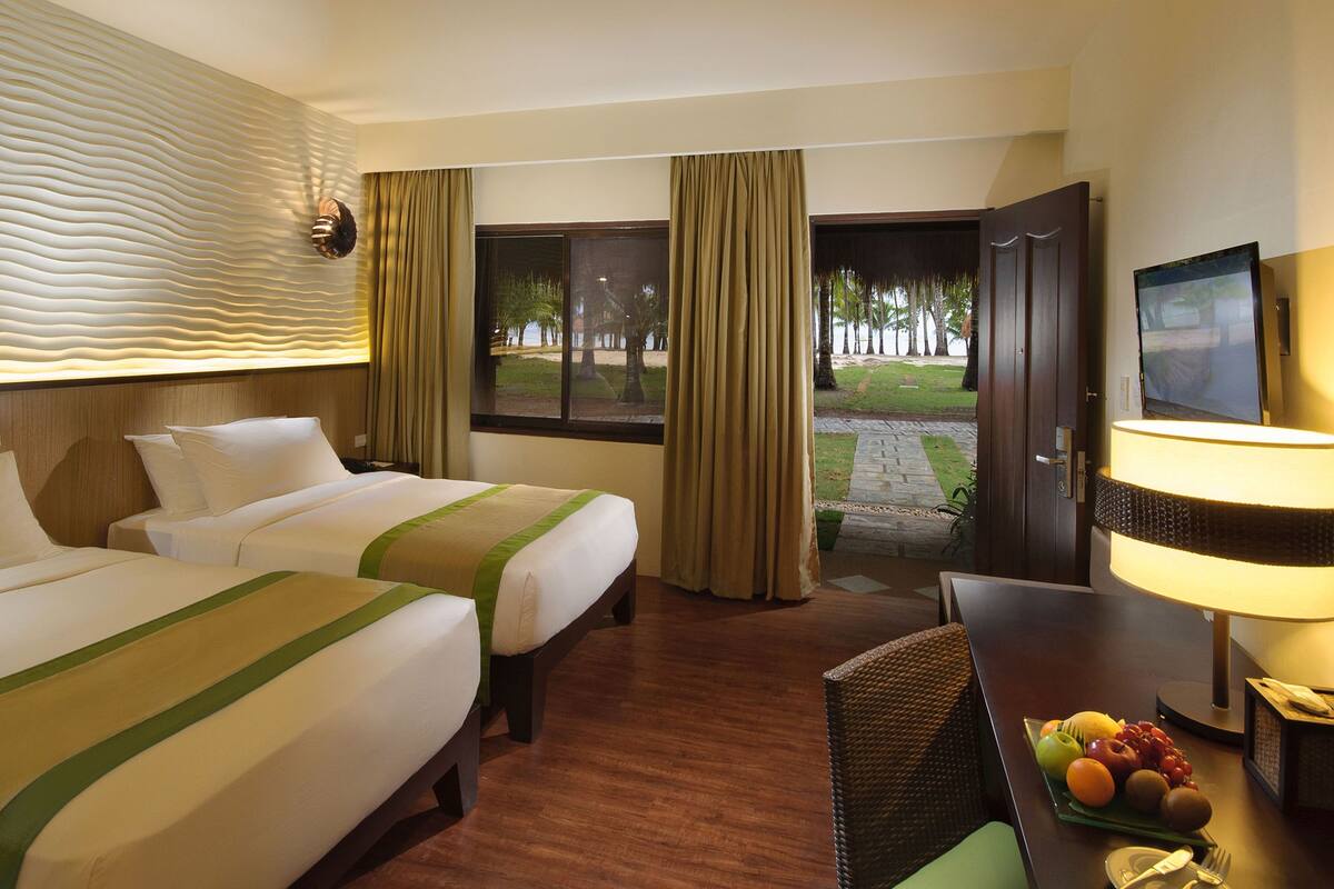 Habitación de hotel con dos camas, un escritorio con lámpara, televisión y vistas a la playa.