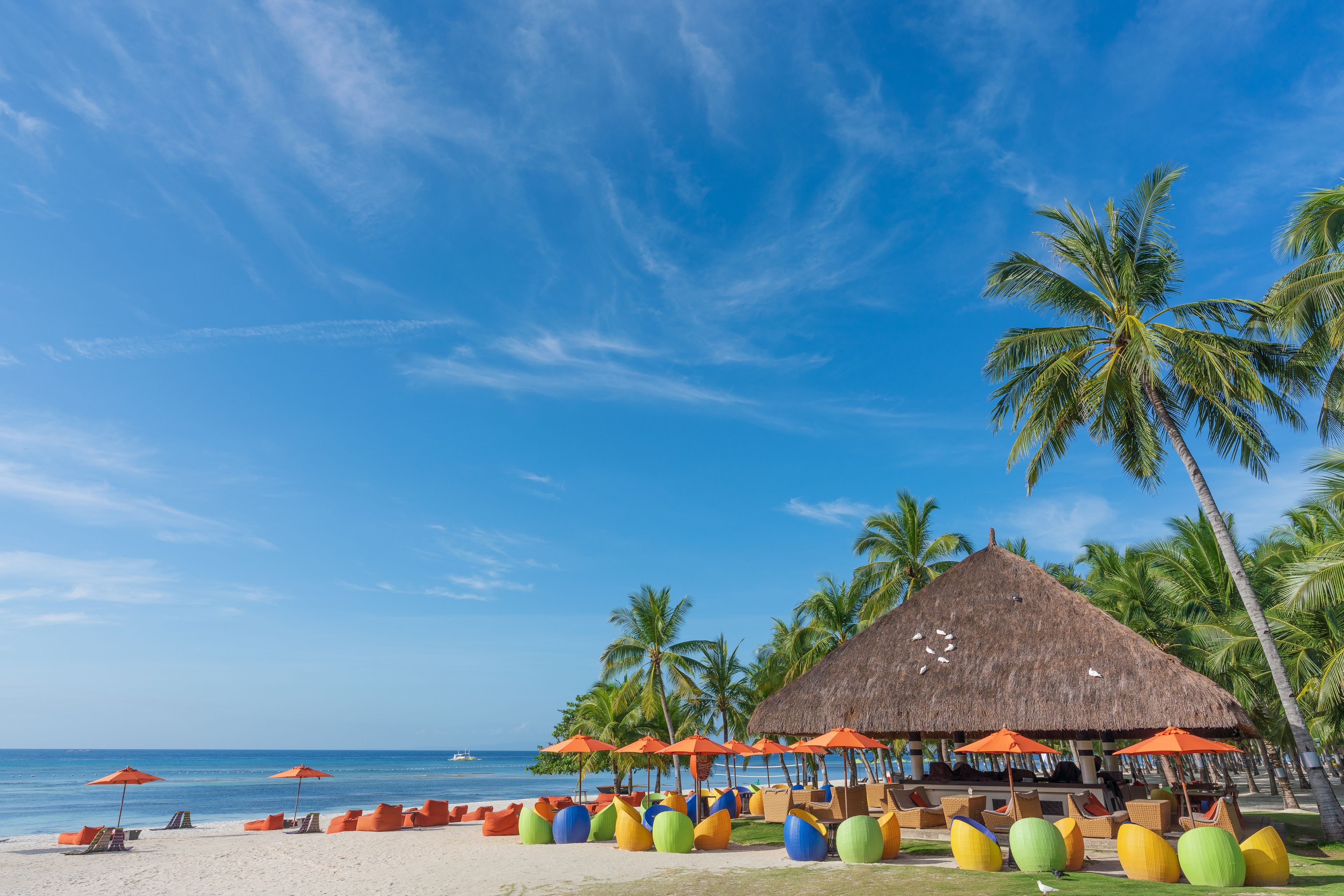 Una playa con una cabaña de techo de paja, coloridos sillones de tumba y palmeras.