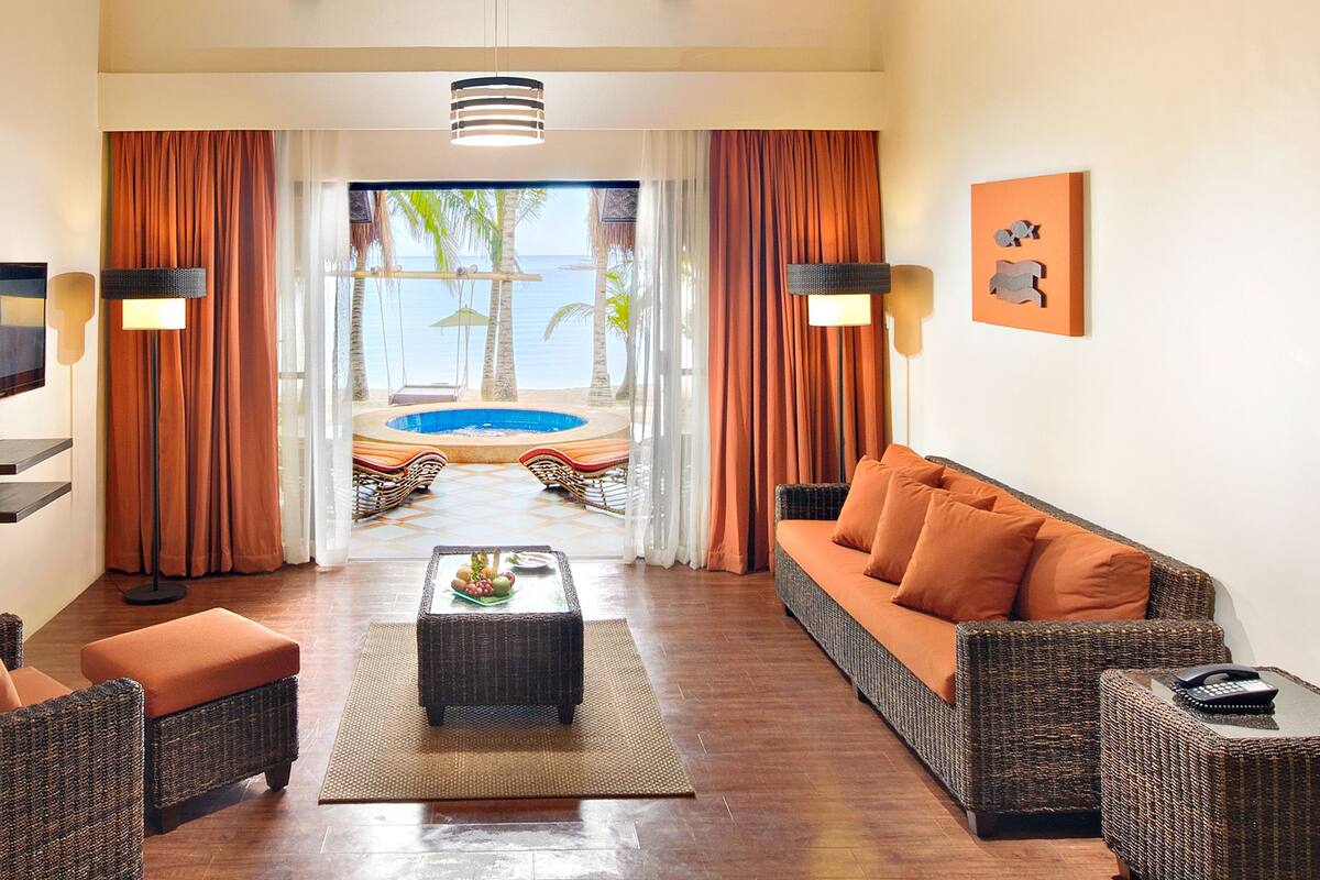 Habitación de hotel con sofá, sillón y una pequeña mesa con frutero. Se ve una piscina y palmeras a través de la ventana.