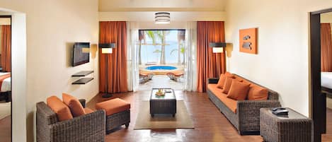 A hotel room with a sofa, armchair, and a small table with a fruit bowl. There is a view of a pool and palm trees through the window.