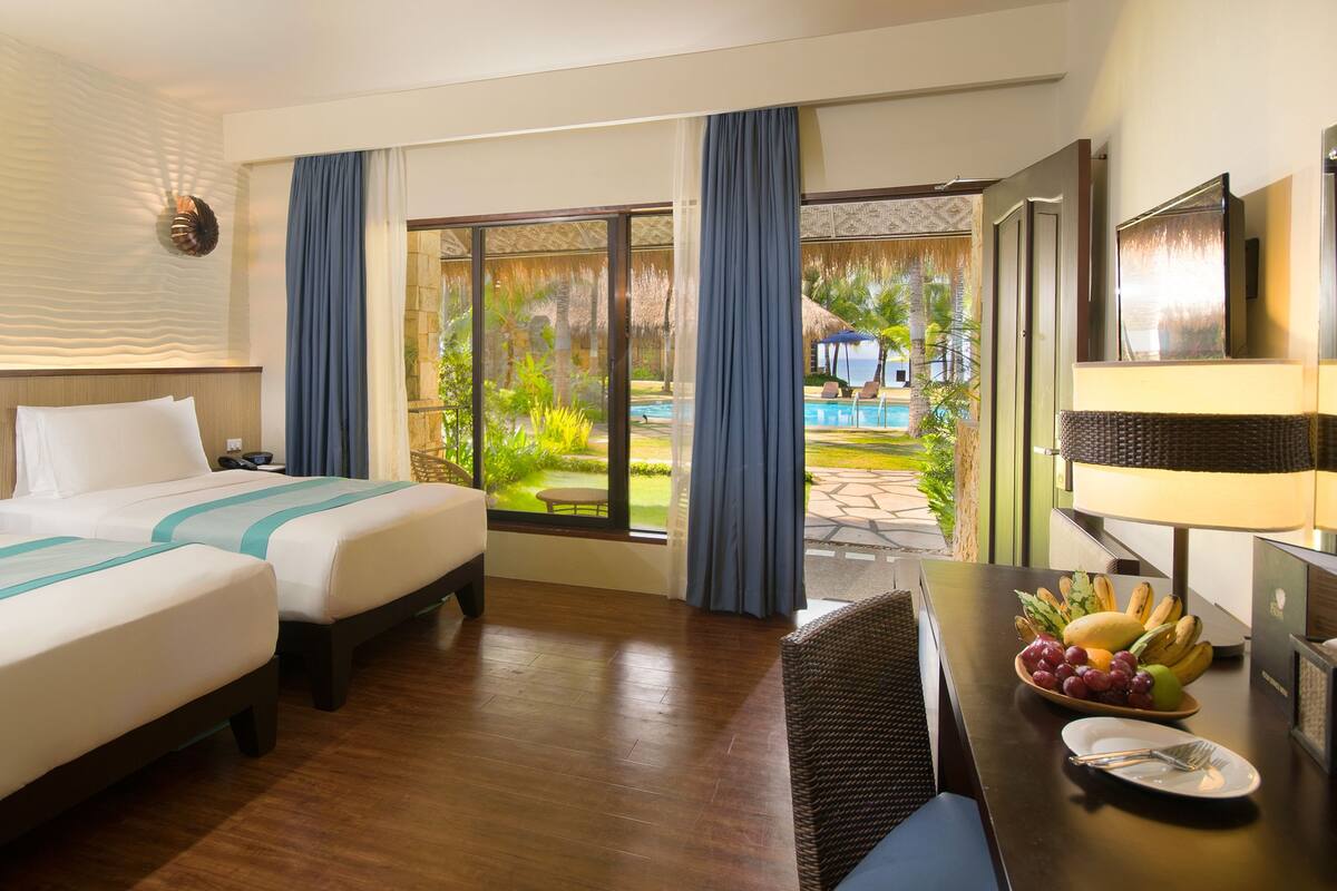 Habitación de hotel con dos camas, una mesa de comedor con fruta, un televisor y vistas a una piscina y jardín.