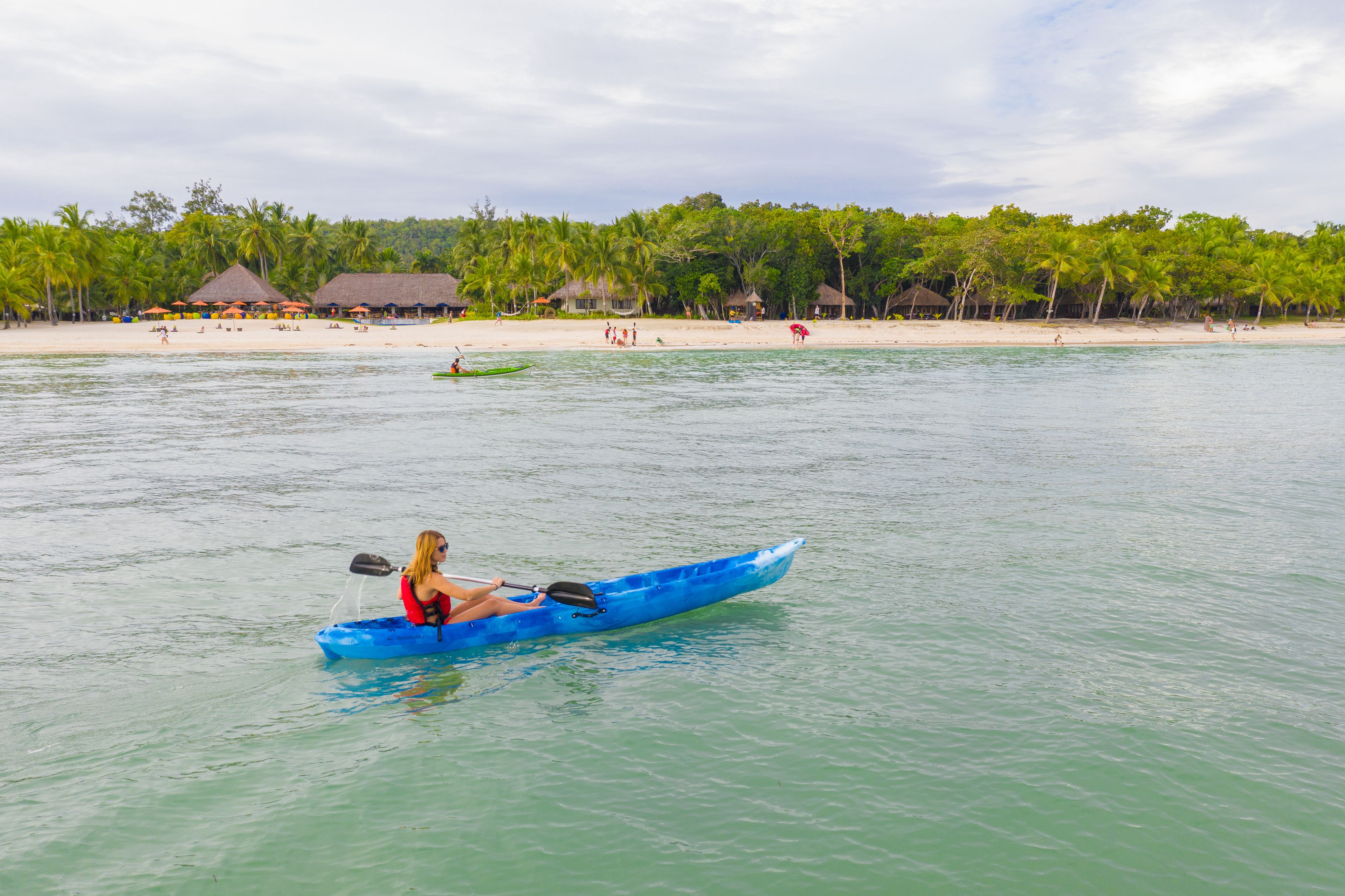 A person kayaking on calm waters with a beach and palm trees in the background.