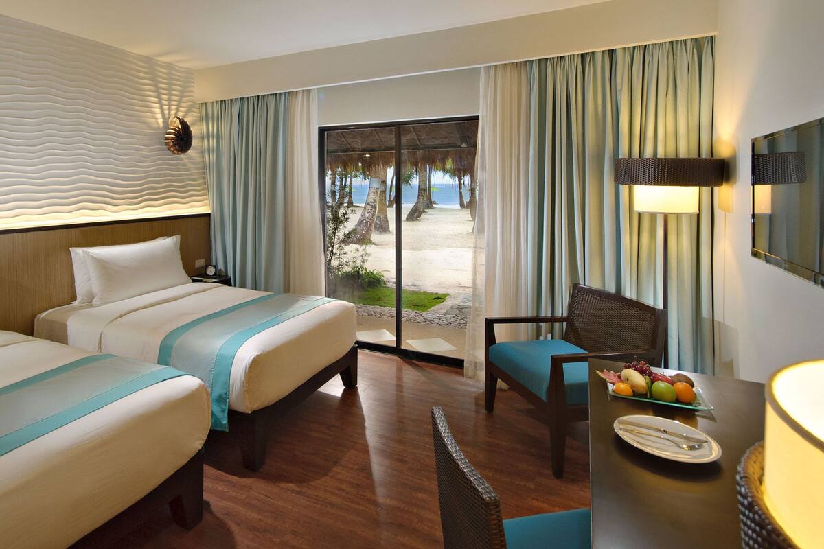 Habitación de hotel con dos camas, un escritorio con silla, vista a la playa y un ventanal con cortinas.